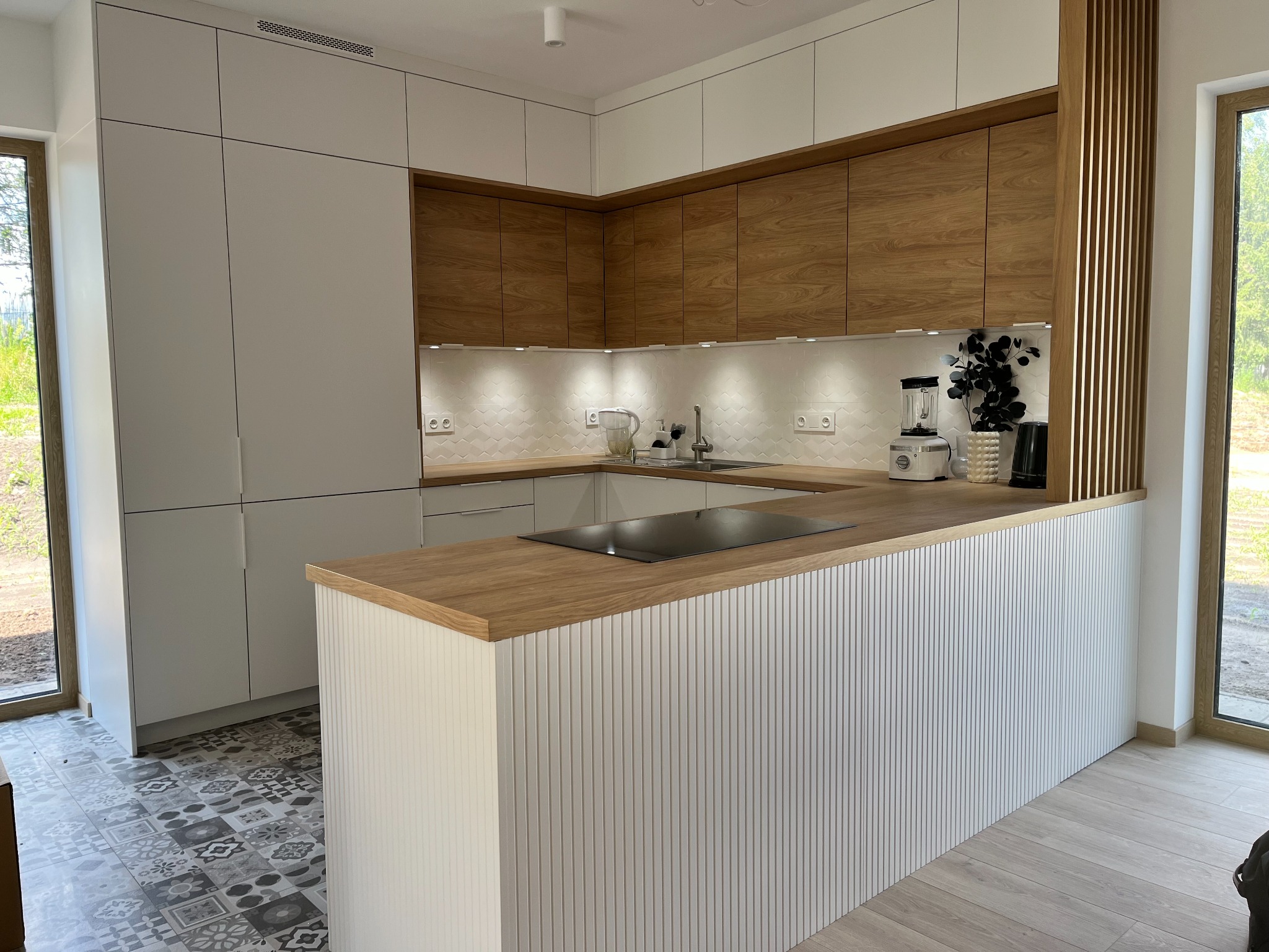 Nowoczesna kuchnia na wymiar z drewnianymi frontami szafek górnych i białymi dolnymi, wyspa z płytą indukcyjną, białe płytki na ścianie i wzorzysta podłoga.