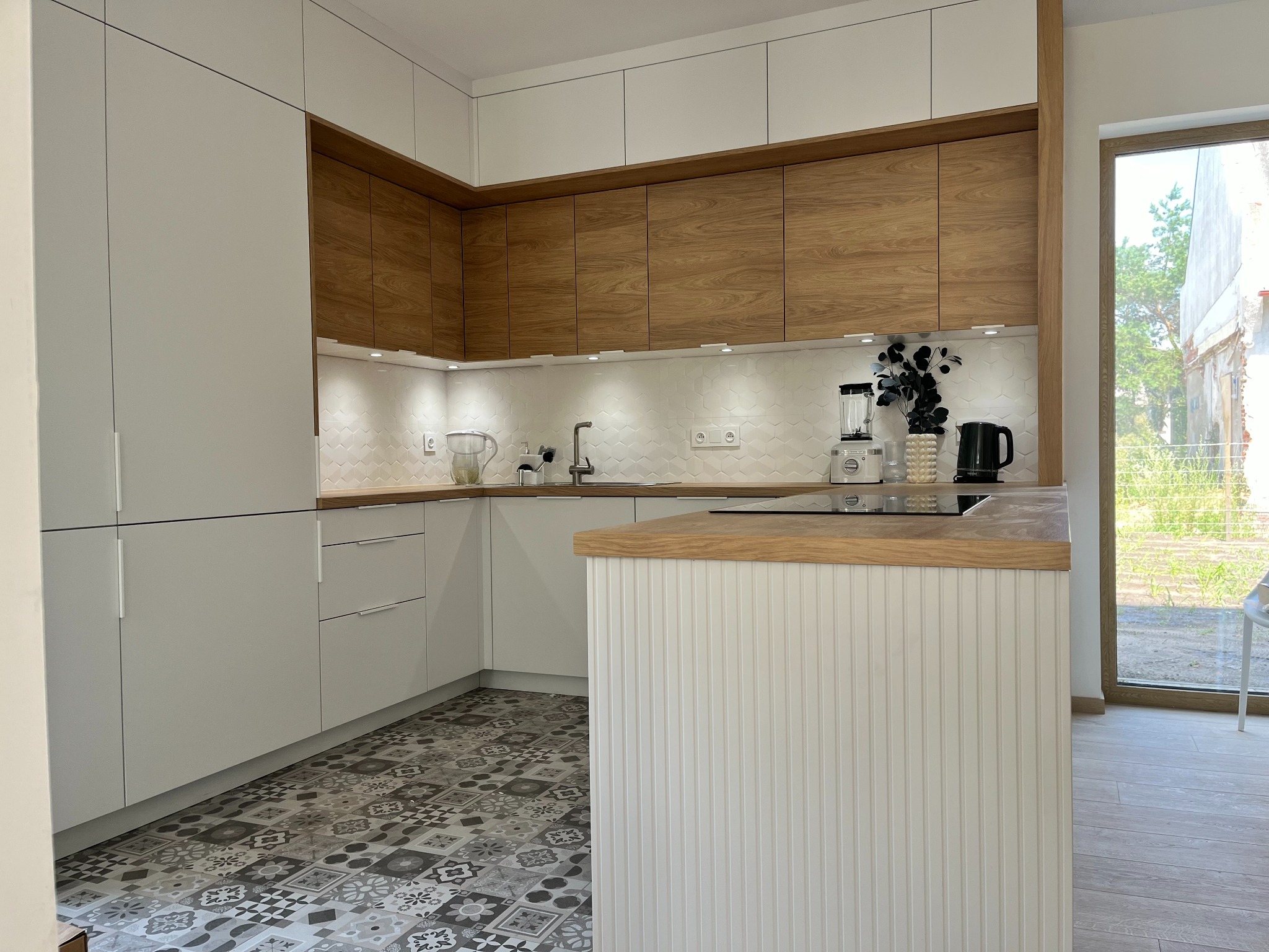 Nowoczesna kuchnia z białymi szafkami i drewnianymi akcentami. Na podłodze wzorzyste płytki. Wyspa kuchenna z blatem z jasnego drewna. Widok na zewnątrz przez okno.