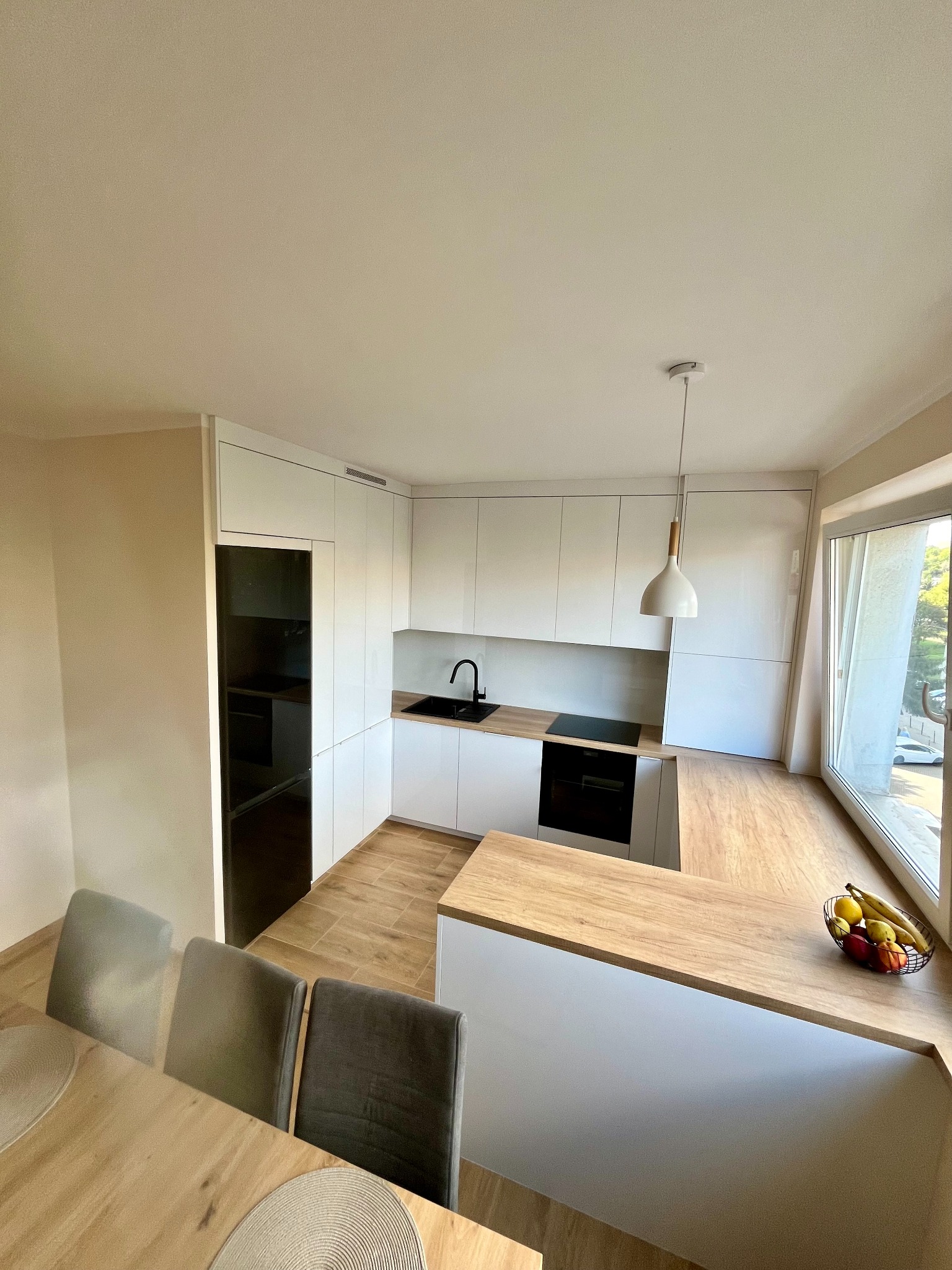 Jasna kuchnia z białymi szafkami i drewnianym blatem, czarny zlew i płyta indukcyjna. Na blacie koszyk z owocami. Widok z jadalni, trzy krzesła przy stole.