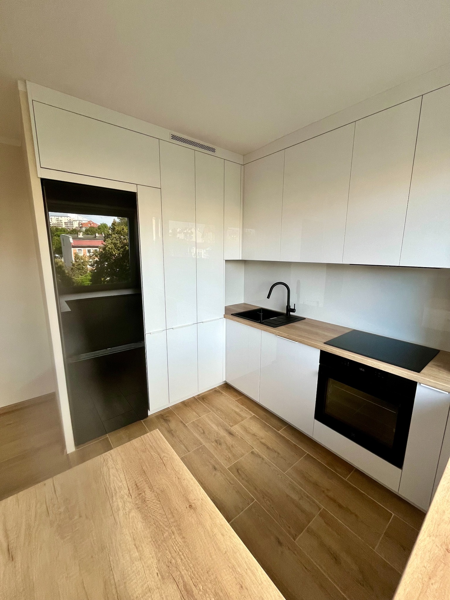 Biała, nowoczesna kuchnia z czarnymi akcentami: zlew, bateria i piekarnik. Widok na zewnątrz przez okno w zabudowie. Drewnopodobne blaty i podłoga.