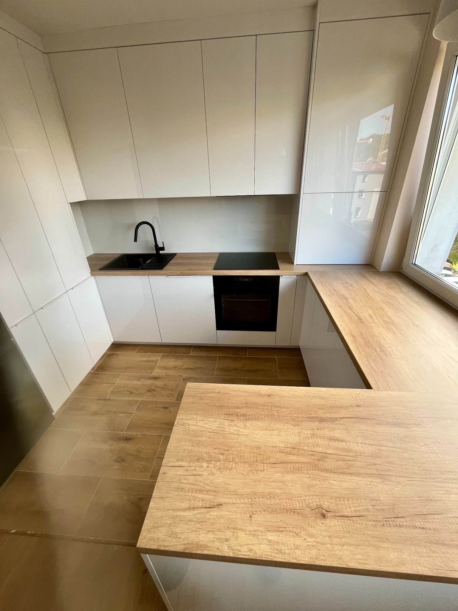 Nowoczesna kuchnia z białymi, lakierowanymi frontami, drewnianym blatem i czarną armaturą. Widok na okno z blatem roboczym i drewnianą podłogą.