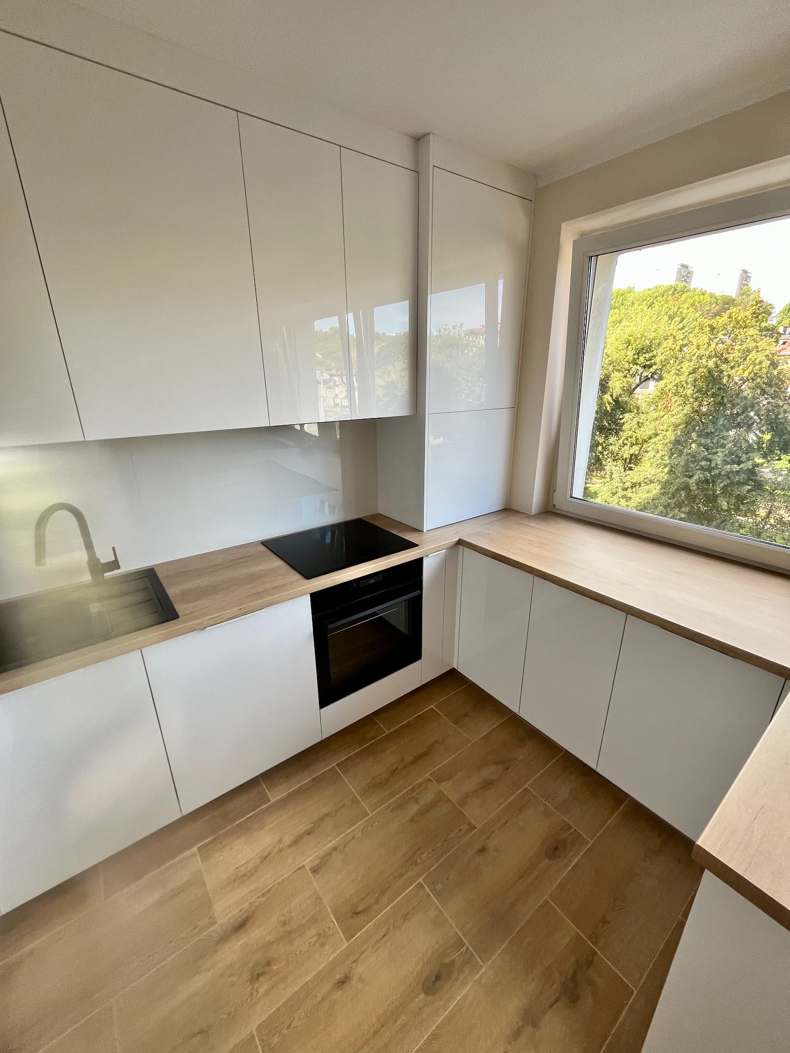 Biała, nowoczesna kuchnia z drewnianym blatem i podłogą. Widok na zieleń za oknem. Minimalistyczny design, błyszczące fronty szafek, czarna płyta indukcyjna i piekarnik.