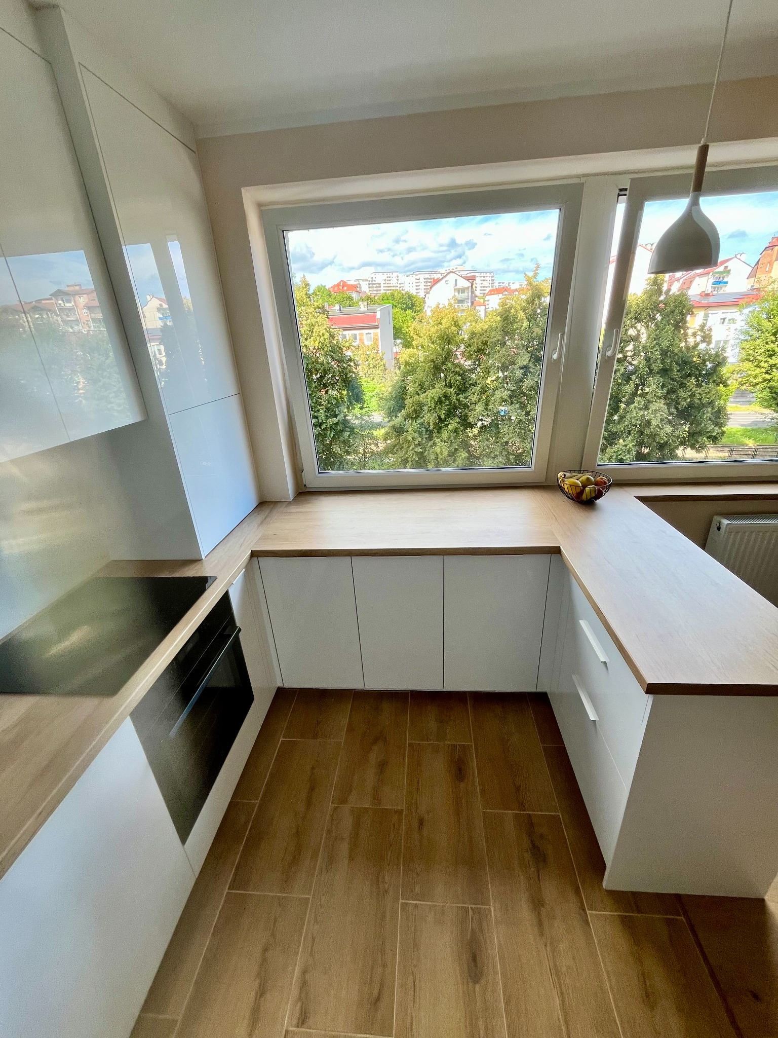 Nowoczesna kuchnia na wymiar z białymi szafkami i drewnianym blatem pod oknem z widokiem na zieleń miejską. Płyta indukcyjna i piekarnik w zabudowie.