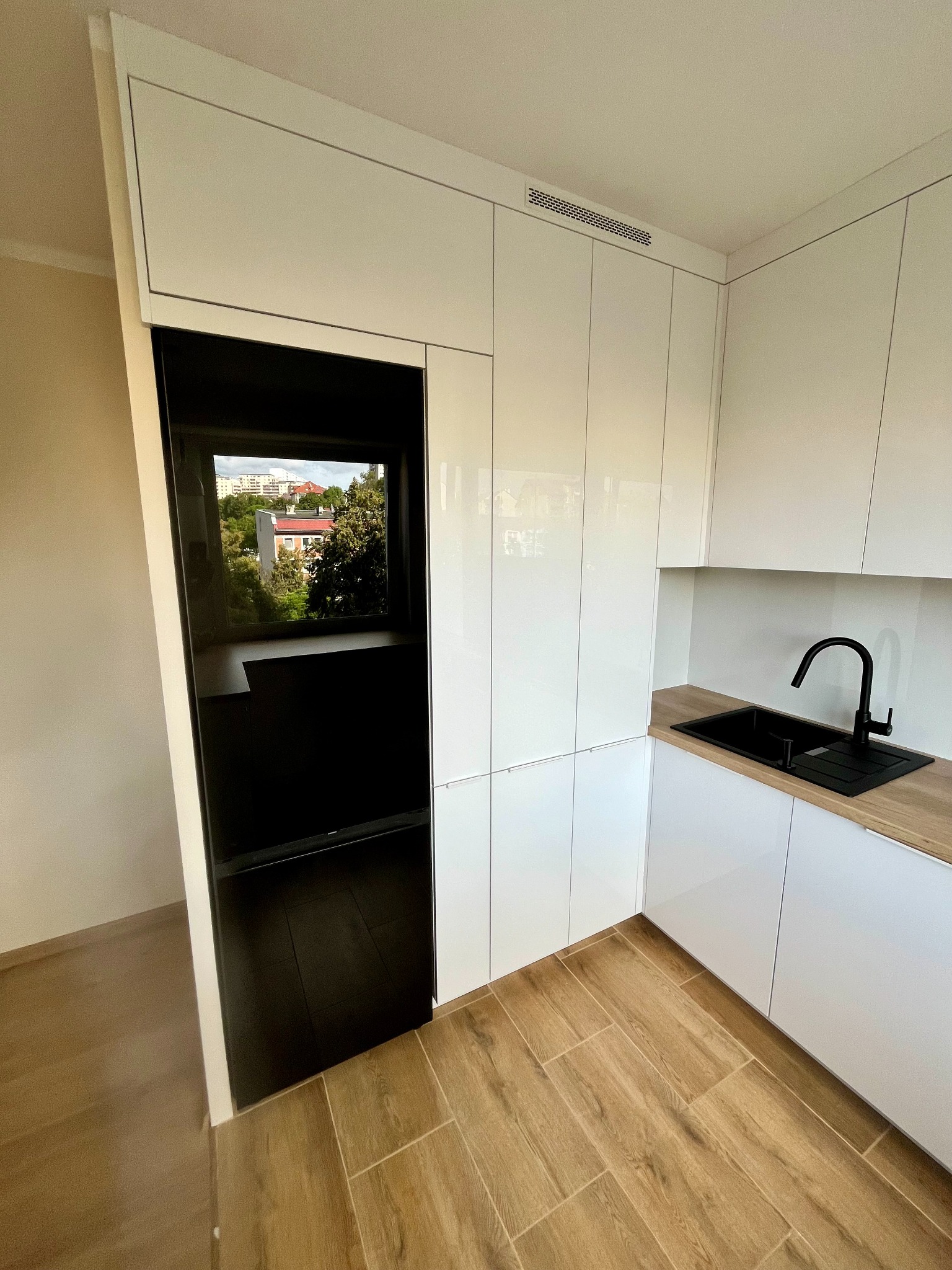 Biała, nowoczesna kuchnia z połyskiem, czarny zlew i bateria, drewniany blat, okno z widokiem na zieleń. Minimalistyczny design, jasne wnętrze, drewniana podłoga.