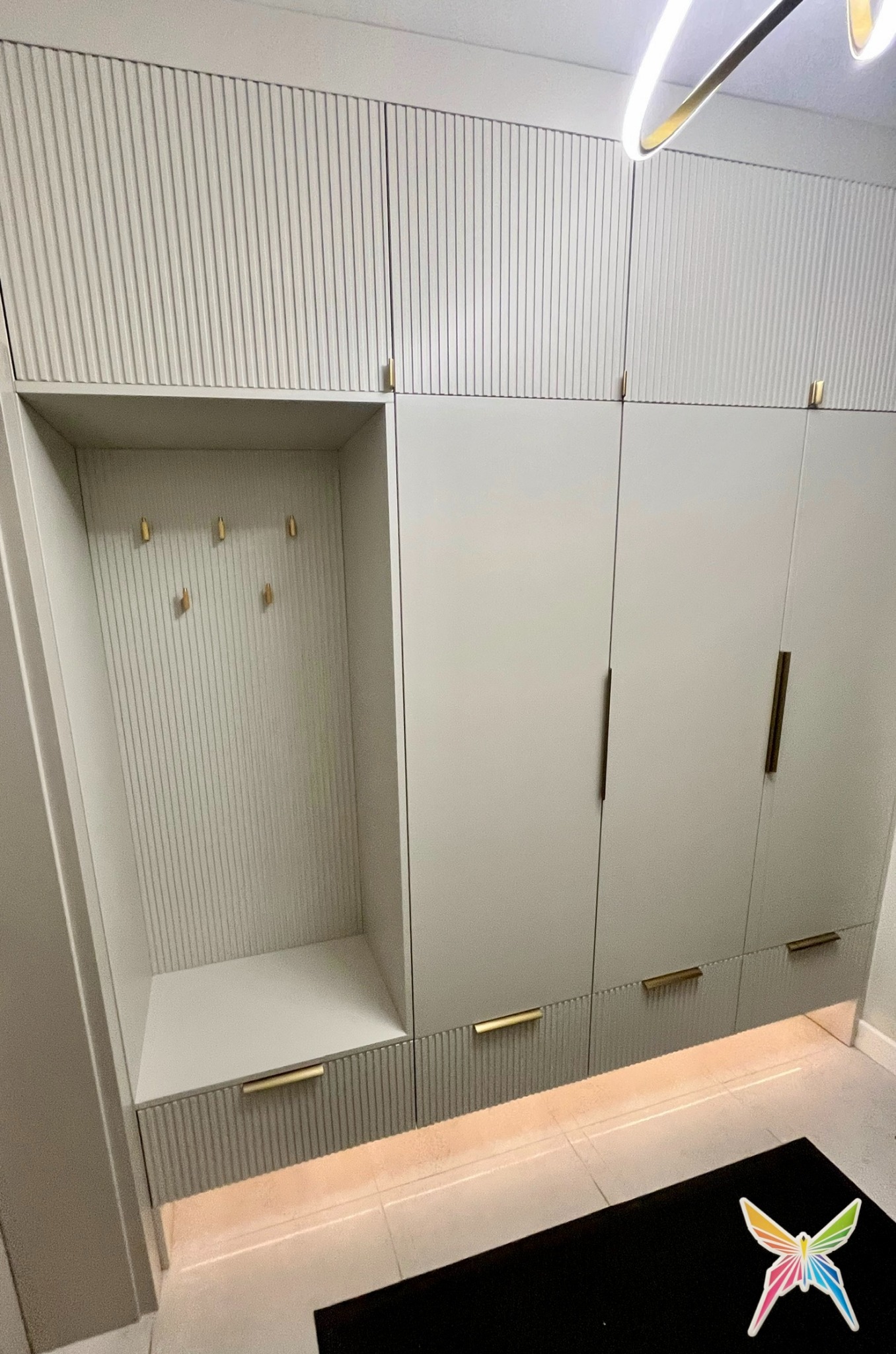 Szara zabudowa meblowa z ryflowanymi frontami i złotymi uchwytami, wnęka z wieszakami, oświetlenie LED przy podłodze, nowoczesny design.