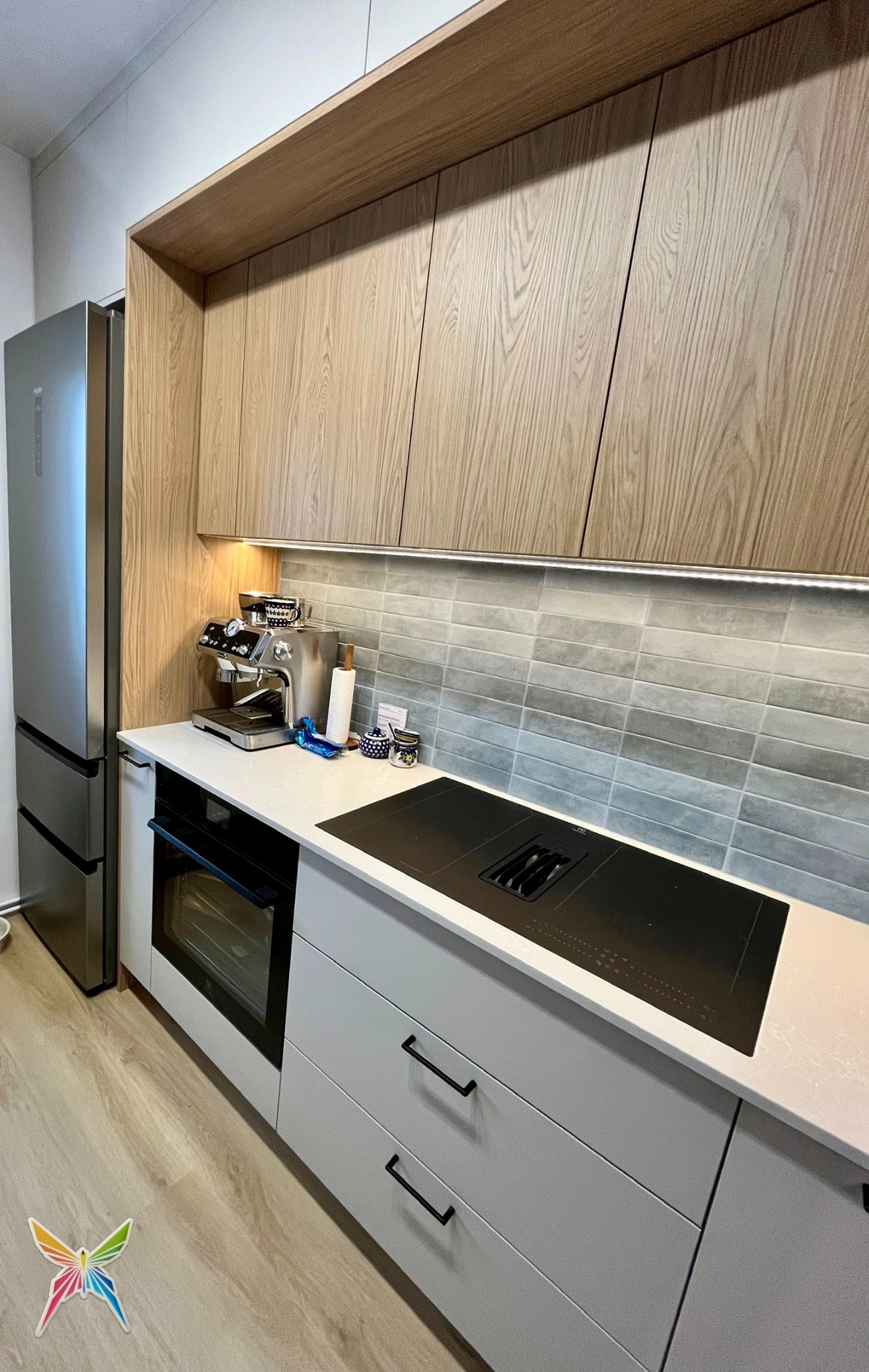 Nowoczesna kuchnia z drewnianymi szafkami górnymi i szarymi dolnymi. Na blacie ekspres do kawy i płyta indukcyjna. Uwagę przykuwa subtelne oświetlenie podszafkowe.