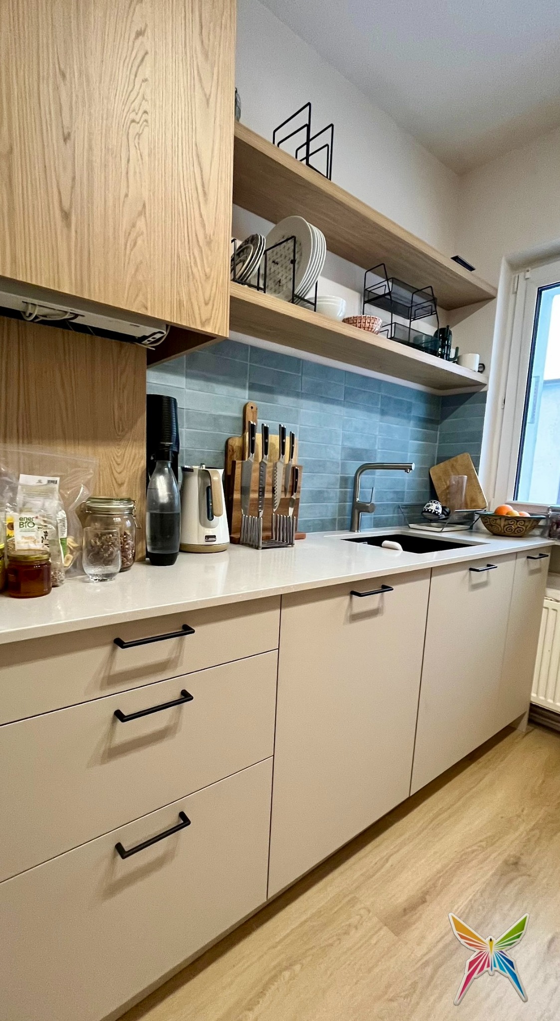 Jasna kuchnia z drewnianymi półkami i błękitną mozaiką. Na blacie akcesoria kuchenne i produkty spożywcze. Minimalistyczne uchwyty szafek.