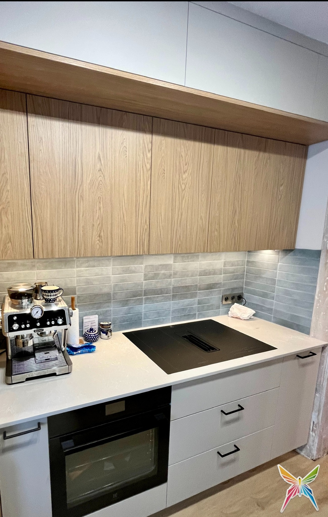 Jasna kuchnia z drewnianymi szafkami wiszącymi i szarymi dolnymi, czarną płytą indukcyjną i ekspresem do kawy na blacie. Minimalistyczny design, płytki w odcieniach szarości.