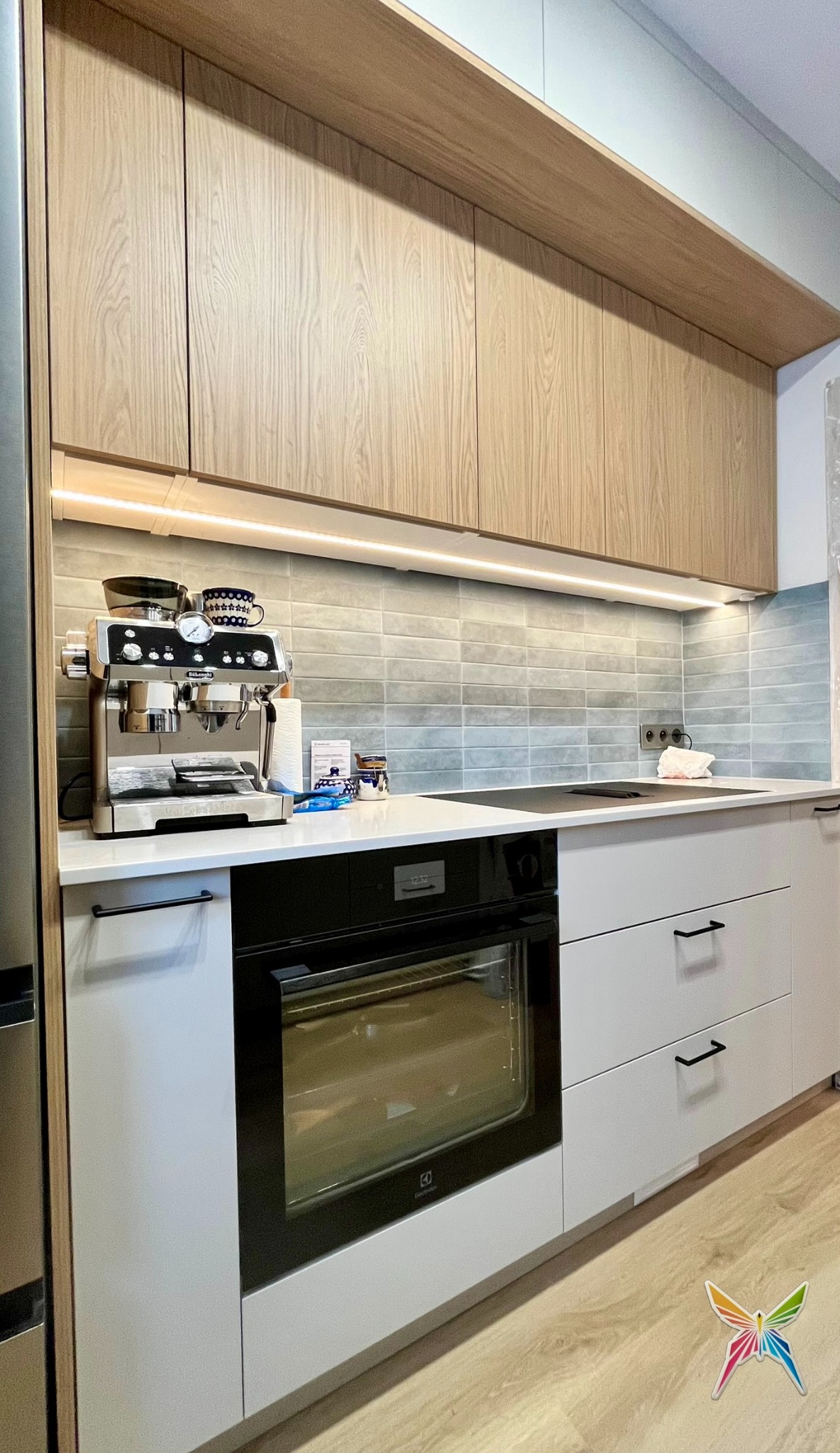 Nowoczesna kuchnia na wymiar z drewnianymi szafkami górnymi, białymi dolnymi, piekarnikiem Electrolux i ekspresem do kawy. Podświetlenie LED pod szafkami.