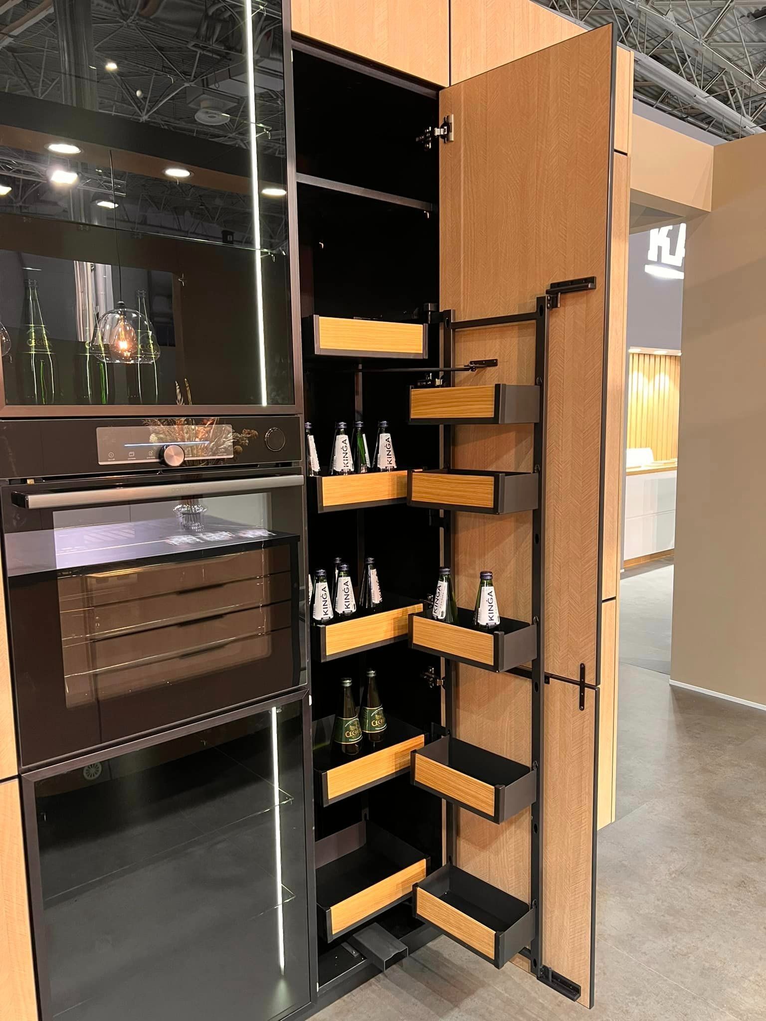 Nowoczesna zabudowa kuchenna z piekarnikiem i systemem cargo z półkami na butelki. Fronty w odcieniach drewna i czerni. Oświetlenie LED wewnątrz szafek.