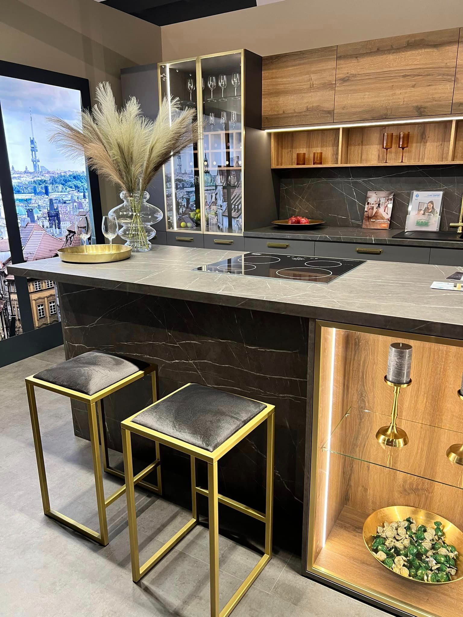 Nowoczesna kuchnia z wyspą, marmurowym blatem i złotymi akcentami. Hoker z szarym siedziskiem i złotą ramą. Podświetlane witryny eksponują dekoracje.