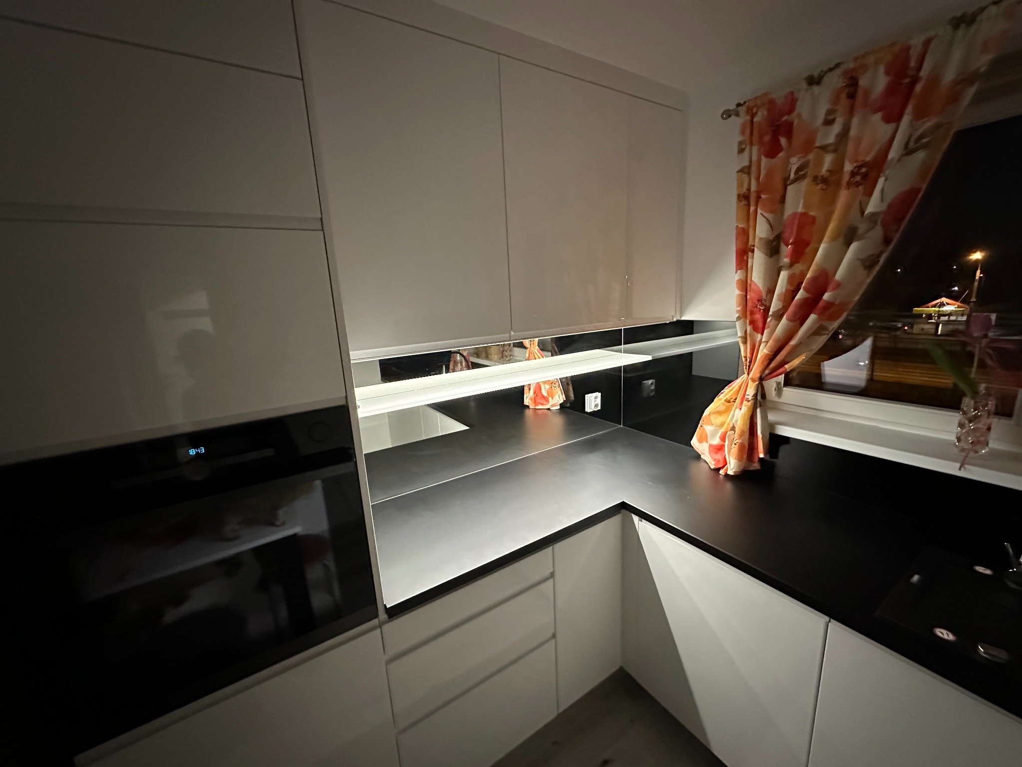 Nowoczesna, biała kuchnia z czarnym blatem, oświetlona wieczornym światłem. Widoczne szafki, piekarnik i okno z kwiecistą zasłoną. Minimalistyczny design.