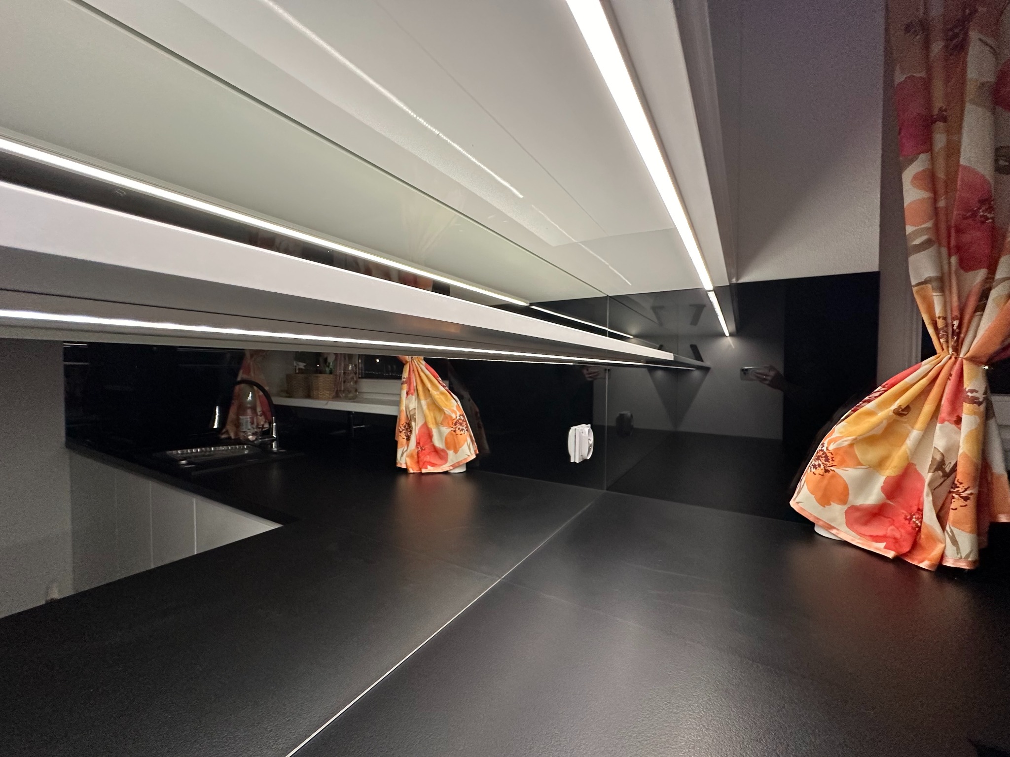 Nowoczesna kuchnia z czarnym blatem i białymi szafkami, oświetlona listwami LED. Widoczne detale wykończenia i zasłona w kwiaty.