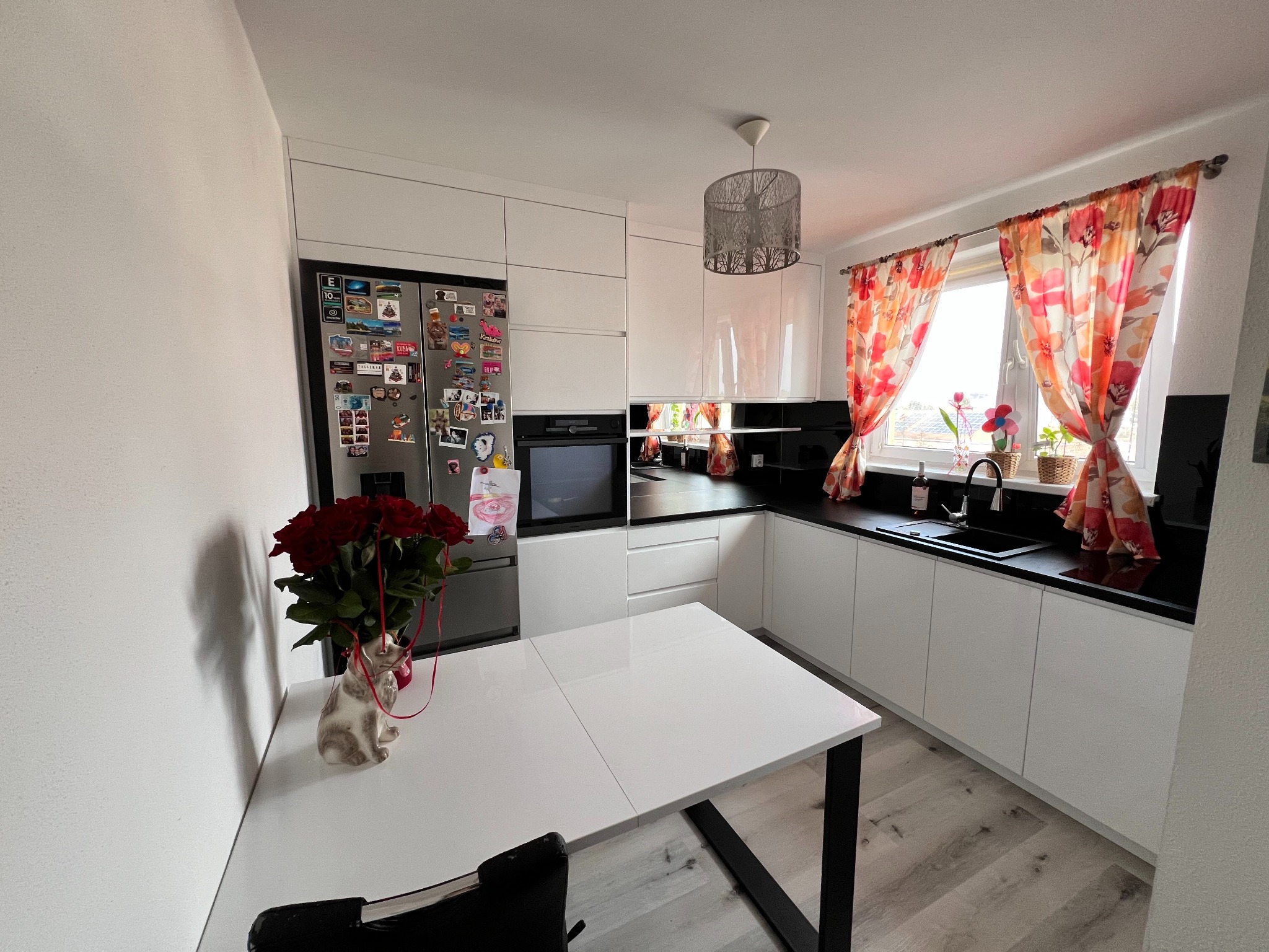 Biała kuchnia na wymiar z czarnym blatem, kwiatami w oknie i bukietem czerwonych róż na stole. Nowoczesny design z połyskiem.