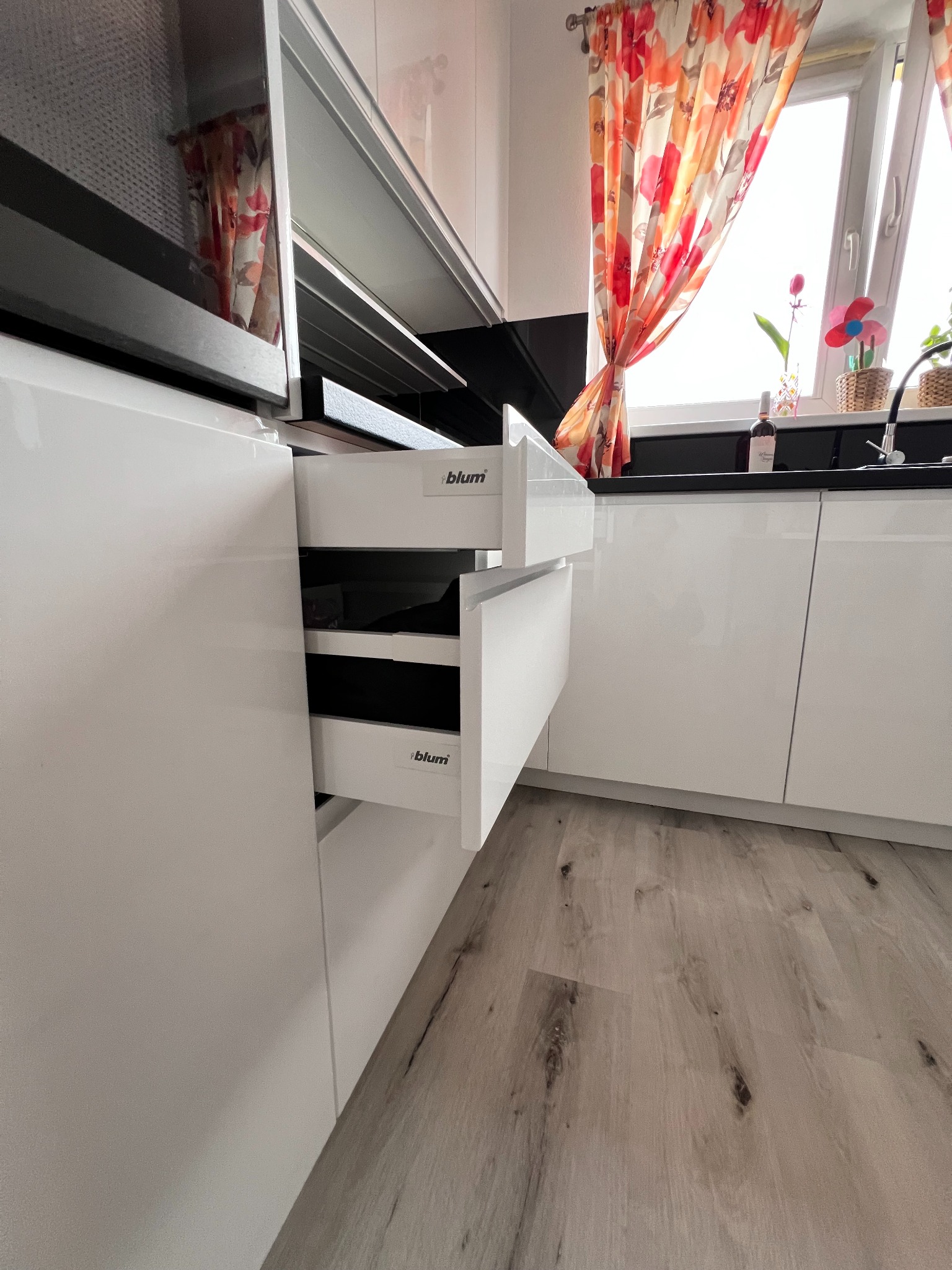 Białe, lakierowane fronty szafek kuchennych z otwartymi szufladami Blum. Na blacie dekoracje okienne i butelka wina. Jasna podłoga z paneli.