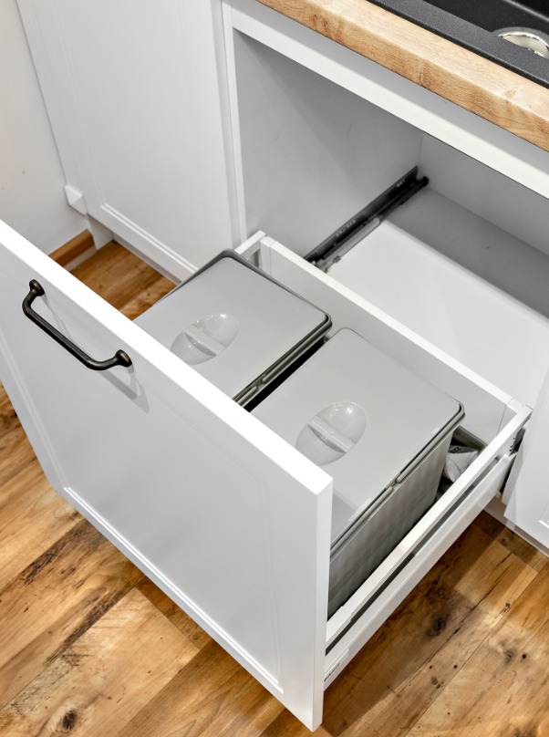Biała szafka kuchenna z wysuwanym koszem na śmieci. Dwa pojemniki z szarymi pokrywami w otwartej szufladzie. Drewniana podłoga i blat. Ciemny uchwyt.