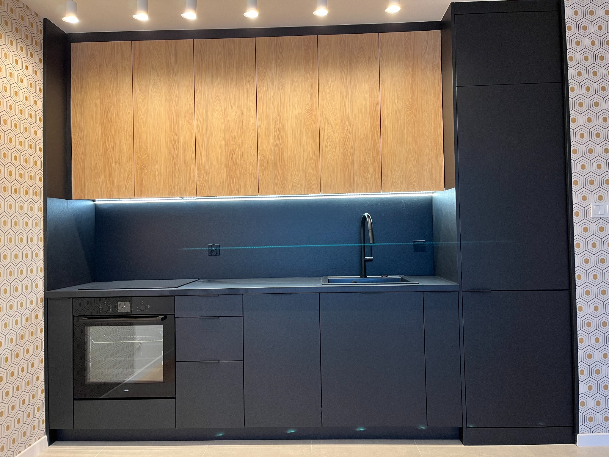 Nowoczesna kuchnia z ciemnymi szafkami i drewnianymi górnymi frontami, oświetleniem LED pod szafkami, z piekarnikiem i zlewozmywakiem. W tle wzorzysta tapeta.