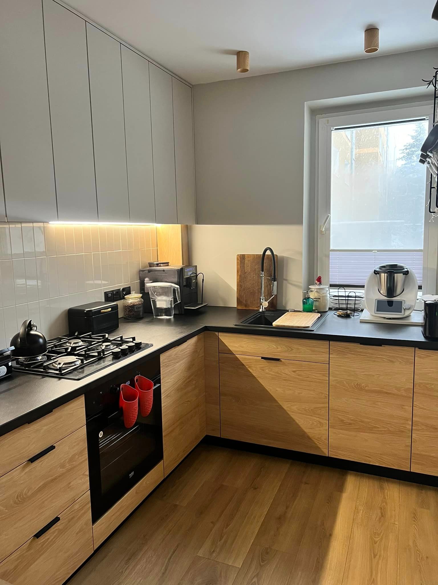 Nowoczesna kuchnia z drewnianymi frontami szafek dolnych i szarymi górnymi, czarny blat, płyta gazowa, piekarnik z czerwonymi rękawicami, okno z roletą.