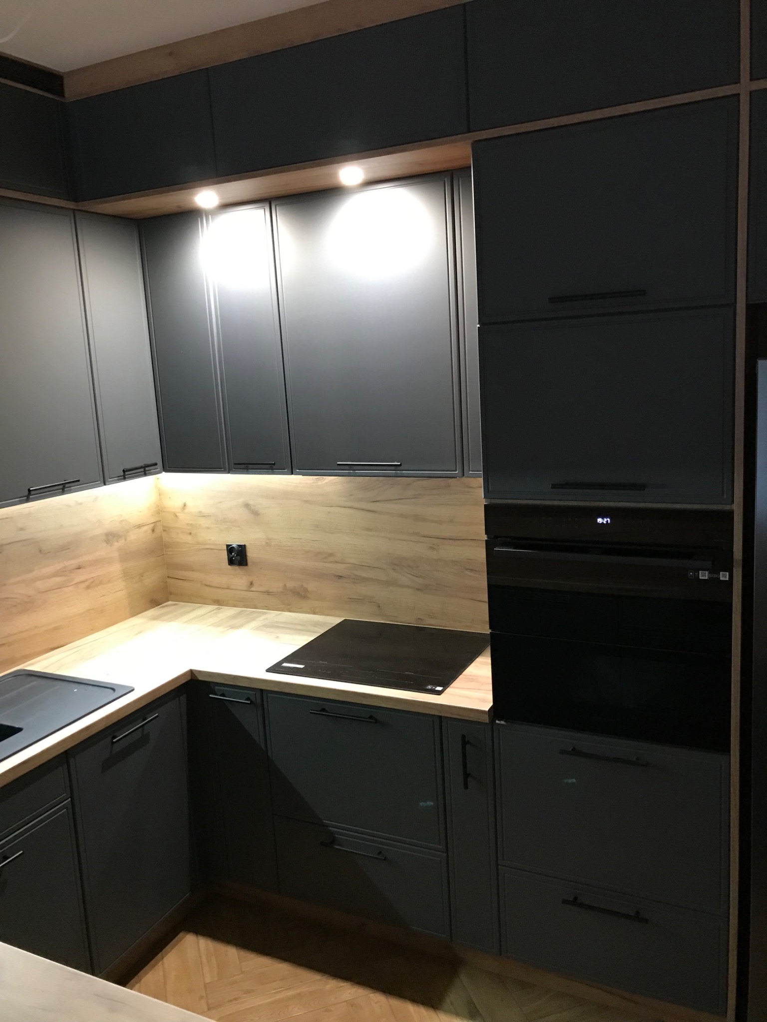 Nowoczesna kuchnia z szarymi matowymi frontami, drewnianym blatem i oświetleniem LED podszafkowym, widoczny piekarnik z wyświetlaczem i płyta indukcyjna.