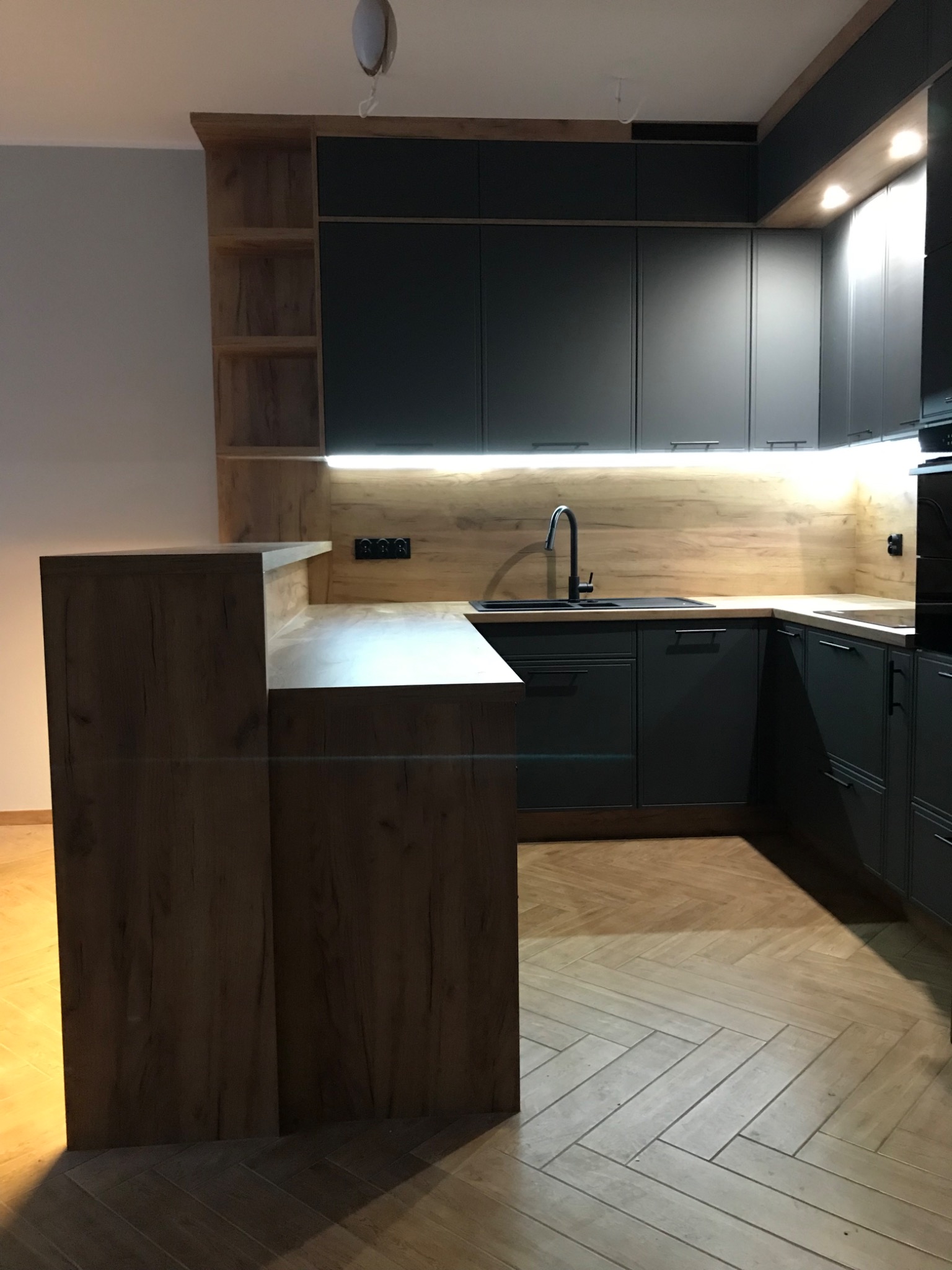 Nowoczesna, szara zabudowa kuchenna z drewnianymi akcentami, oświetleniem LED podszafkowym i podłogą ułożoną w jodełkę.