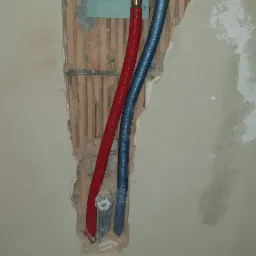 Wnęka w ścianie z wystającymi rurami: czerwona i niebieska, podłączone do zaworu, widoczny fragment wiertarki Milwaukee na podłodze, surowy stan wykończenia.