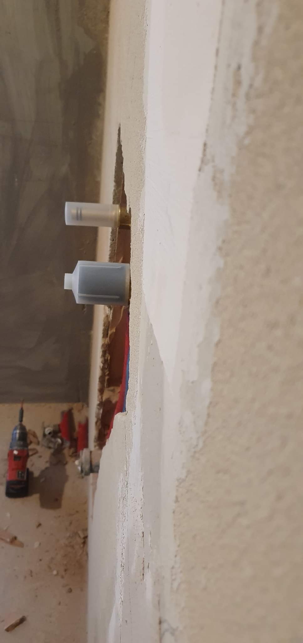 Wystające z otynkowanej ściany końcówki rur wodnych z założonymi plastikowymi zaślepkami, w tle widoczne narzędzia i fragment instalacji.