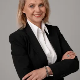 Portret uśmiechniętej blondynki w czarnym garniturze i białej koszuli, z rękami skrzyżowanymi na piersiach, prezentującej srebrną bransoletkę i pierścionek z turkusowym oczkiem.