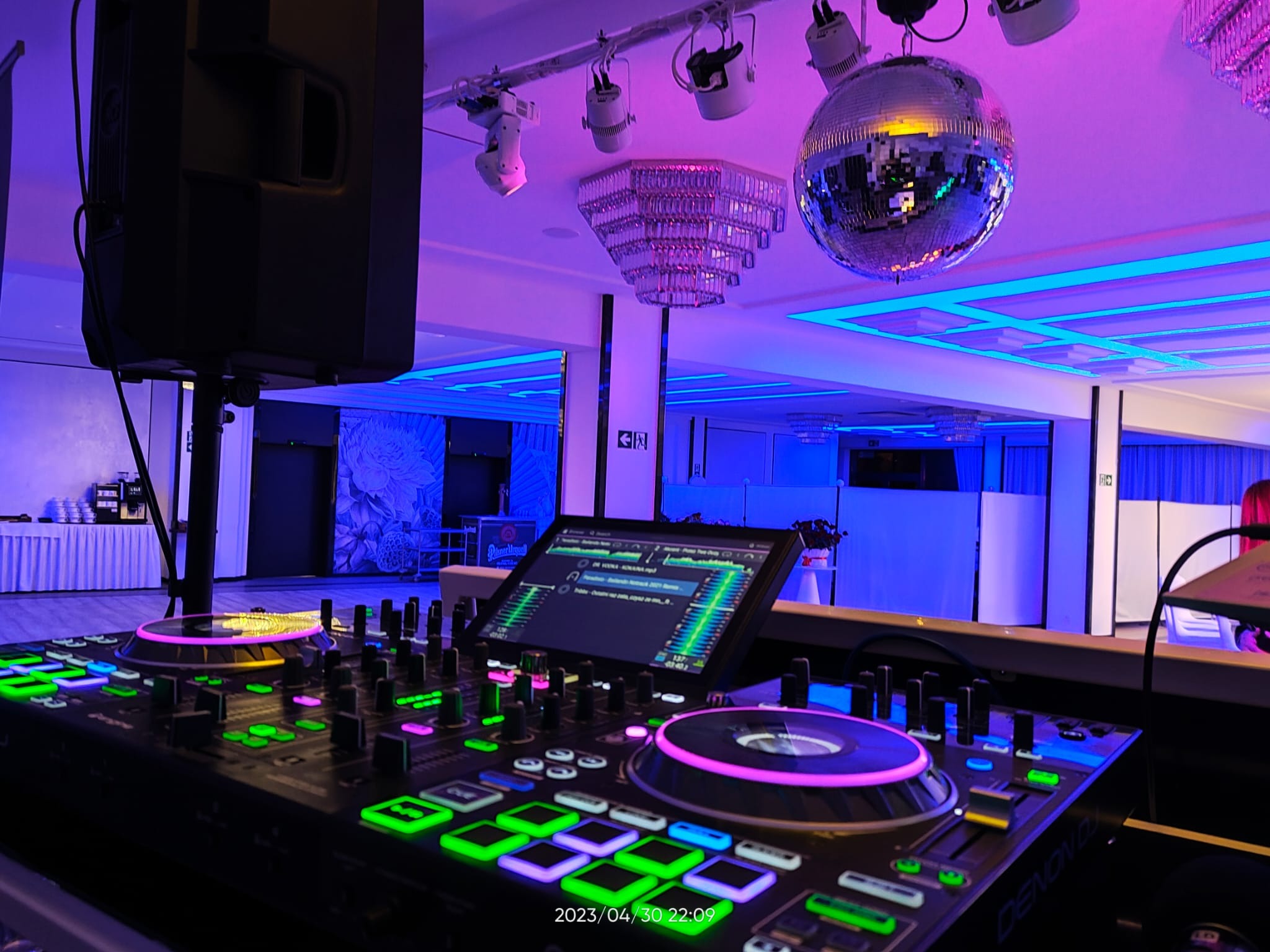 Profesjonalny kontroler DJ Denon z podświetlanymi elementami, ustawiony w sali bankietowej z kryształowym żyrandolem i dyskotekową kulą, oświetlony fioletowo-niebieskim światłem.