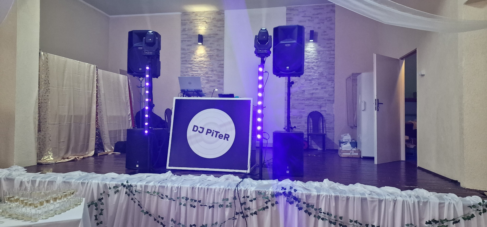Scena z nagłośnieniem, oświetleniem i stanowiskiem DJ-a z logo DJ PiTeR, przygotowana na imprezę, z kieliszkami i dekoracją stołu w pierwszym planie.