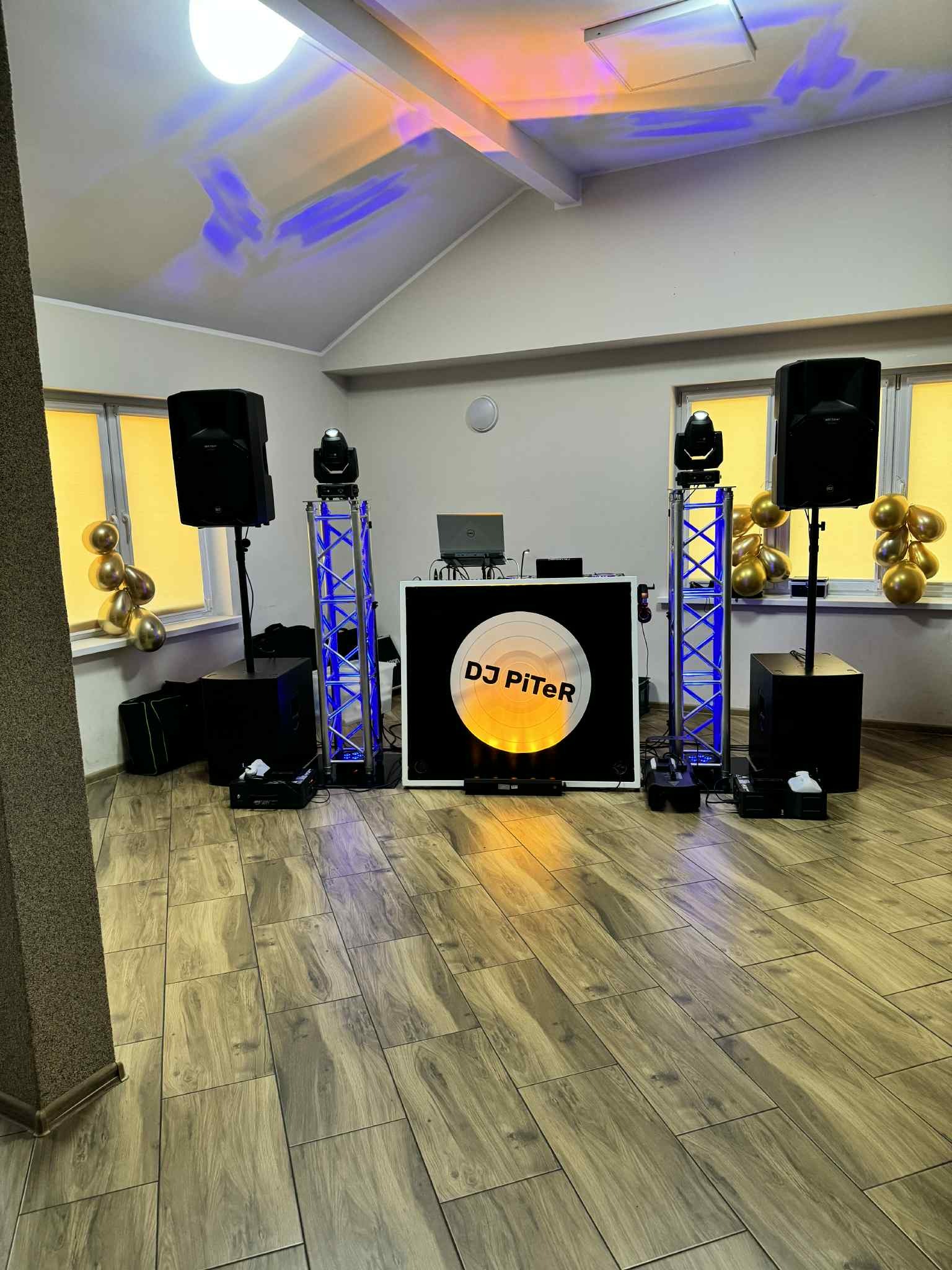 Rozstawiony sprzęt DJ'ski z kolumnami, oświetleniem i stanowiskiem z napisem 'DJ PiTeR', gotowy do występu na parkiecie. W tle okna z balonami.