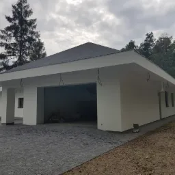 Nowoczesny, parterowy budynek z płaskim dachem i garażem, wykończony na zewnątrz, z kostką brukową na podjeździe, otoczony zielenią. Widoczne wystające kable instalacji elektrycznej.