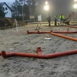Wykonane przyłącze kanalizacyjne z rur PCV na placu budowy, w tle ekipa budowlana z mini koparką i dom jednorodzinny.