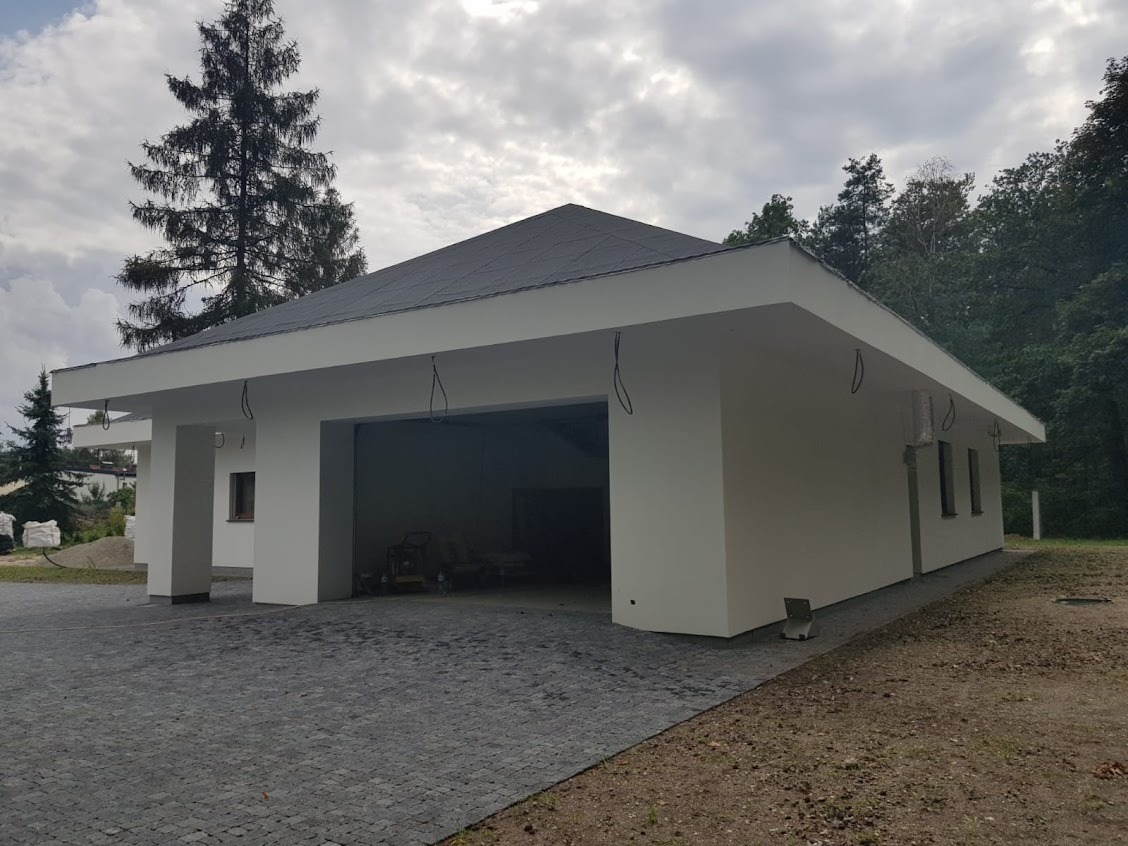 Nowoczesny, parterowy budynek z płaskim dachem i garażem, wykończony na zewnątrz, z kostką brukową na podjeździe, otoczony zielenią. Widoczne wystające kable instalacji elektrycznej.