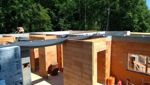 Budowa domu z pustaków ceramicznych; widoczne ściany działowe, otwory okienne i drzwiowe, oraz materiały izolacyjne; w tle zalesiony teren.