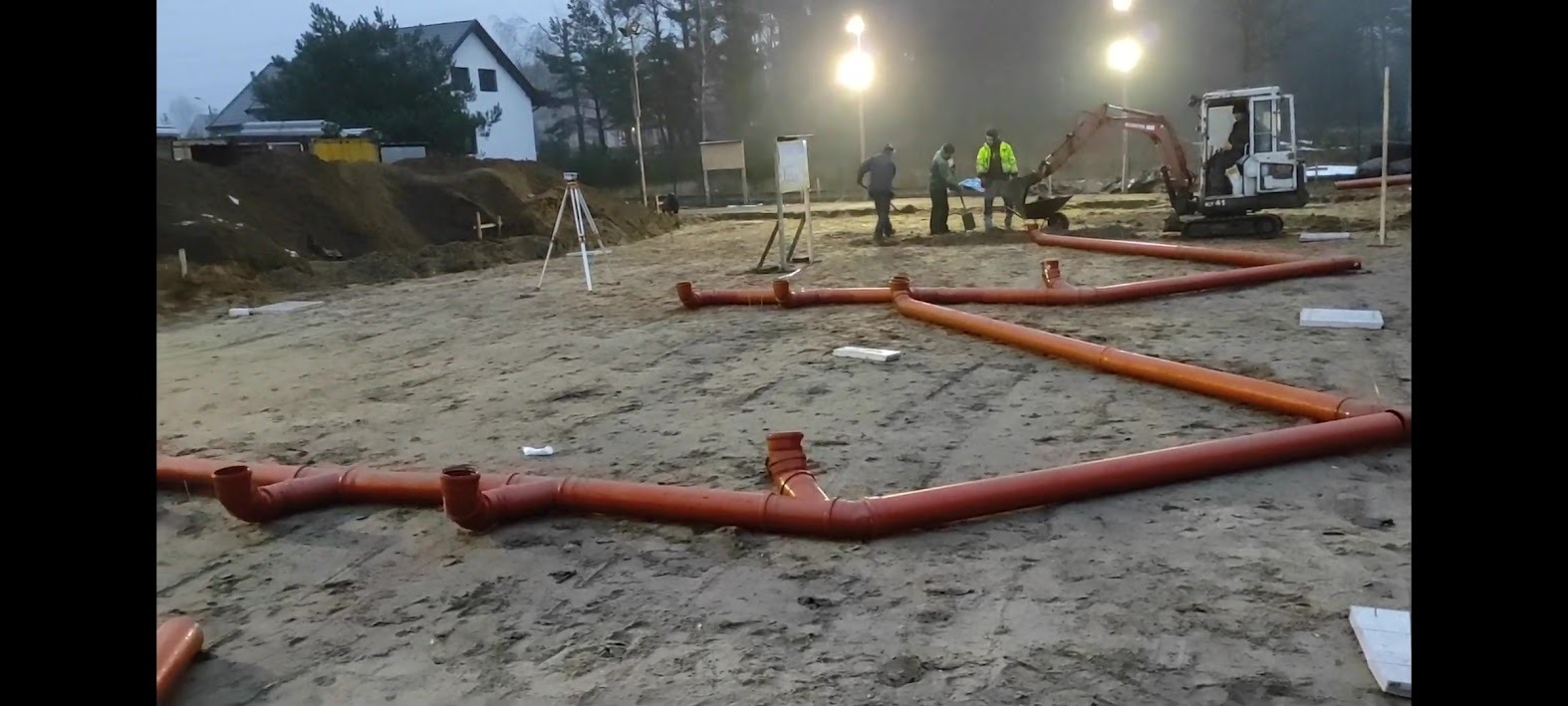 Wykonane przyłącze kanalizacyjne z rur PCV na placu budowy, w tle ekipa budowlana z mini koparką i dom jednorodzinny.
