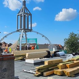 Imperium Dach - Wysoka konstrukcja na dachu w trakcie budowy, widoczne drewniane elementy, materiały izolacyjne i pracownicy w oddali, błękitne niebo z chmurami w tle.