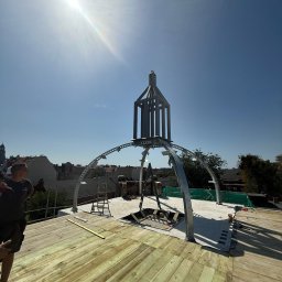 Imperium Dach - Konstrukcja metalowa na dachu, prawdopodobnie element dekoracyjny lub podstawa pod kopułę. Widoczna drewniana podłoga dachu w trakcie budowy i panorama miasta w tle.