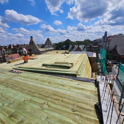 Imperium Dach - Montaż drewnianej konstrukcji dachu na tle błękitnego nieba z obłokami. Widoczni dekarze podczas pracy. Ujęcie z góry, ukazujące postęp prac.