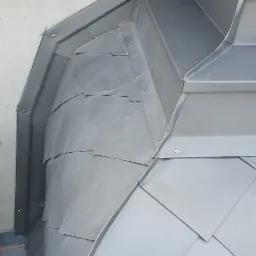 Szczegółowe ujęcie obróbki blacharskiej na dachu zabytkowej kamienicy, z widocznymi elementami dekoracyjnymi elewacji i metalowymi płytkami ułożonymi w unikalny wzór.