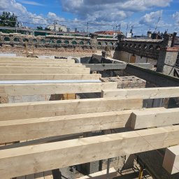 Imperium Dach - Wysoki kąt: Nowa więźba dachowa z jasnego drewna na tle starego, ceglanego budynku z widocznymi elementami zabezpieczeń i błękitnego nieba z chmurami.