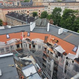 Imperium Dach - Widok z góry na budynek w trakcie renowacji. Część dachu pokryta czerwoną dachówką, część czarnym materiałem. Widoczne rusztowania i pracownicy. W tle zabudowa miejska.
