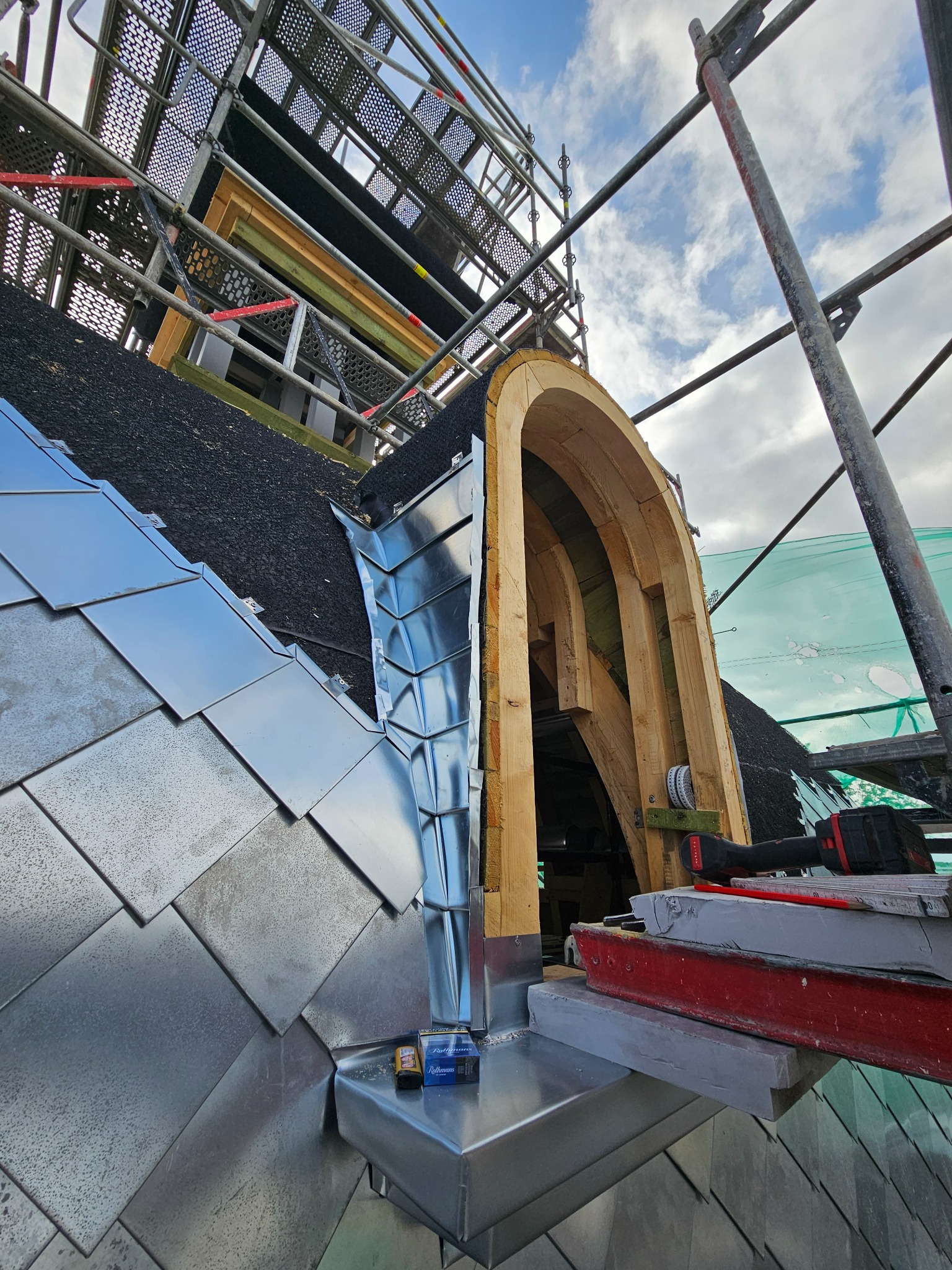 Metalowe płytki na dachu z drewnianą konstrukcją okna dachowego w trakcie montażu. Widoczne rusztowanie i narzędzia pracy.