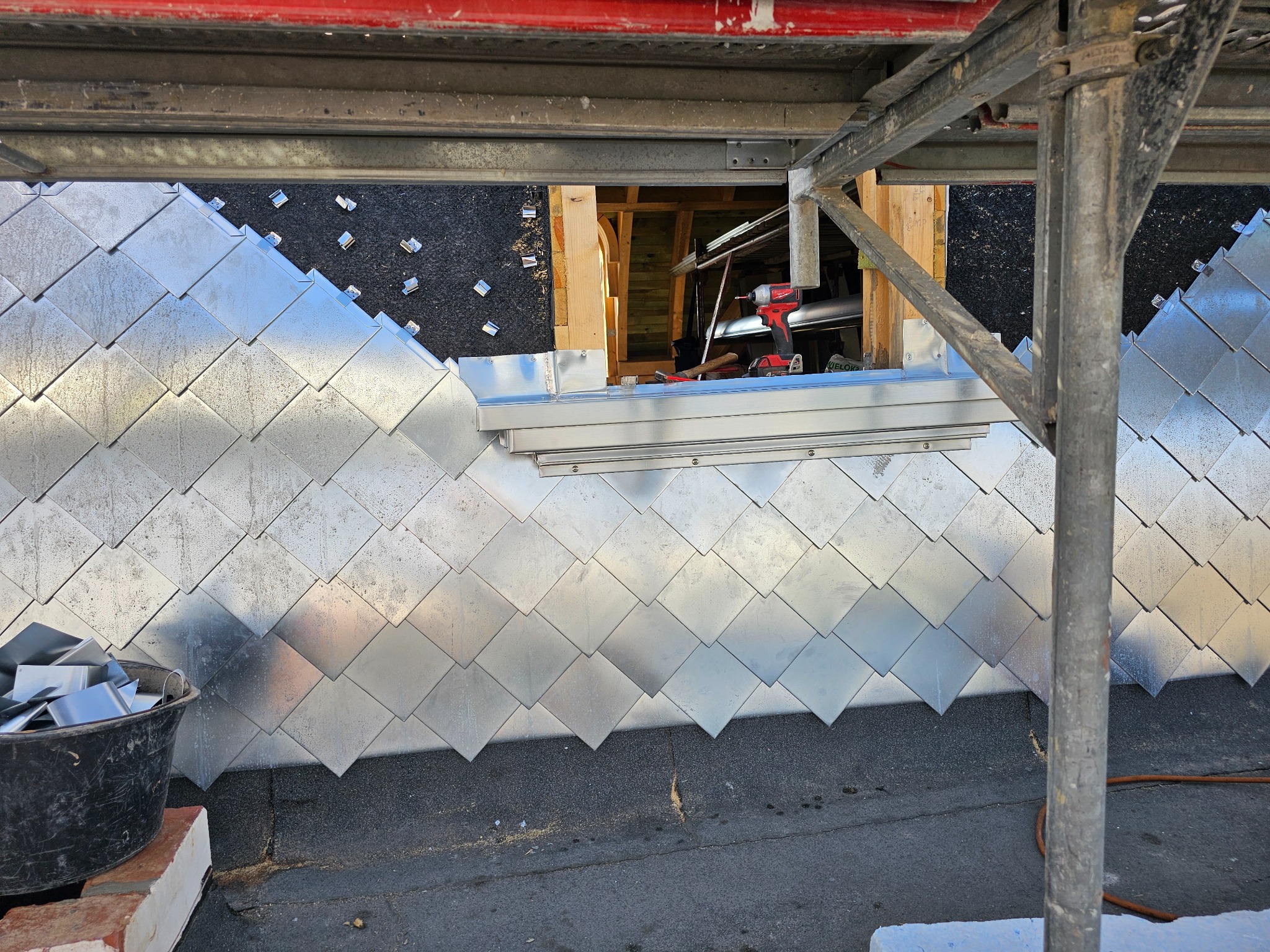 Montaż srebrnych, romboidalnych elementów na dachu, widoczne rusztowanie i narzędzia, etap prac. Nietypowe krycie dachu z blachy w trakcie realizacji.