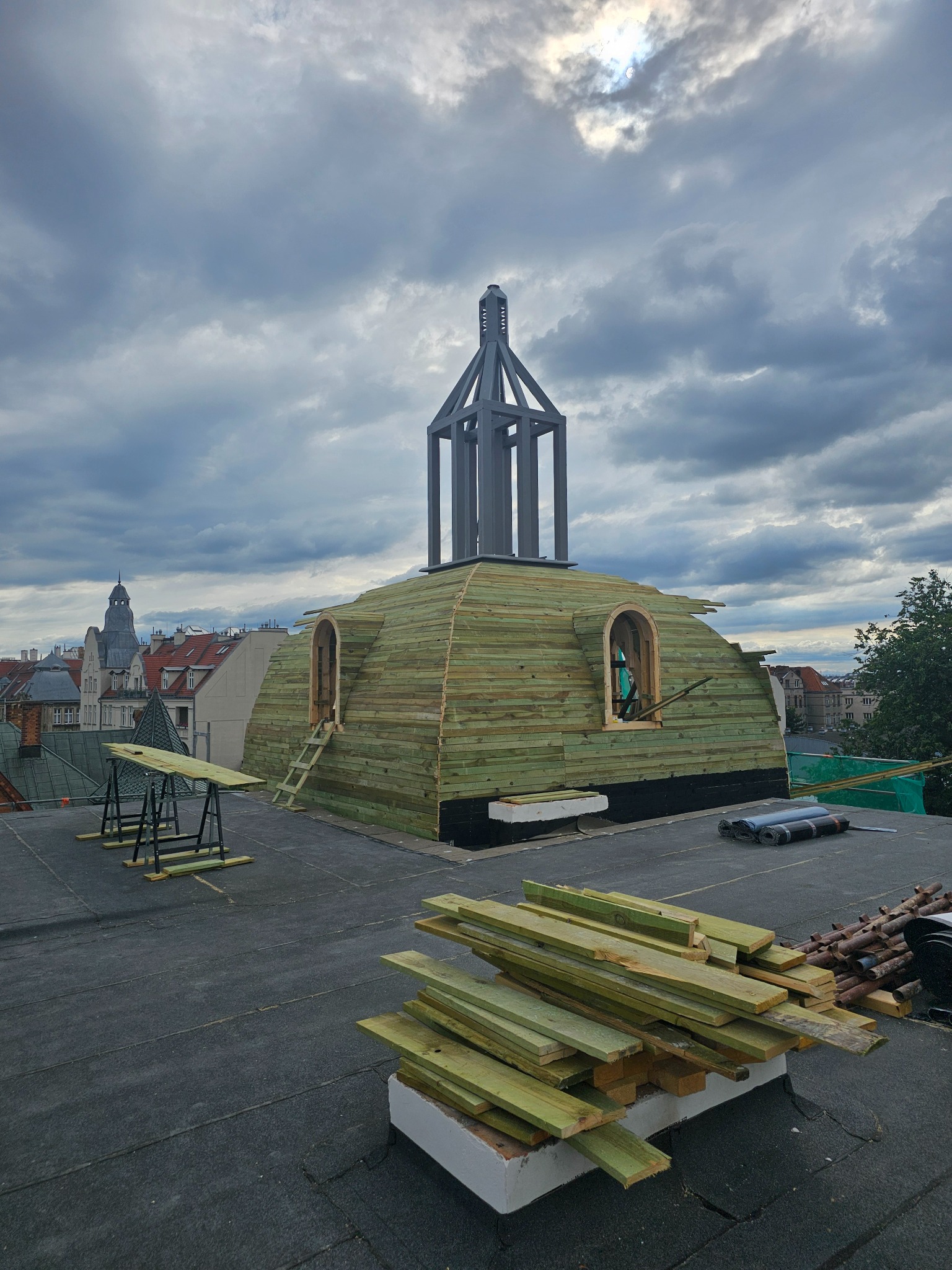 Prace na dachu budynku: drewniana konstrukcja z oknami w trakcie budowy, widoczne deski i narzędzia, w tle zabudowa miejska pod pochmurnym niebem.