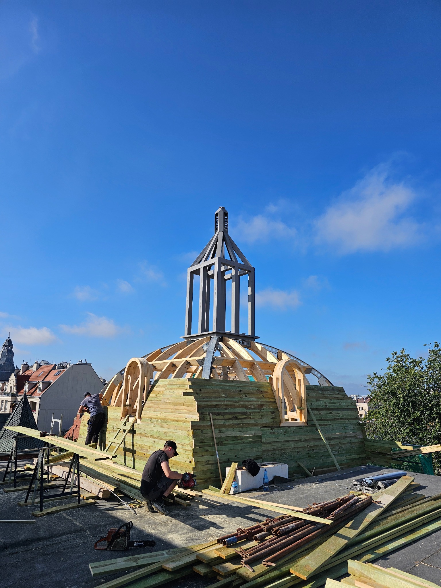 Budowa kopuły na dachu budynku. Dwóch mężczyzn pracuje przy montażu drewnianej konstrukcji. W tle błękitne niebo i zabudowania miejskie. Widoczne narzędzia pracy.
