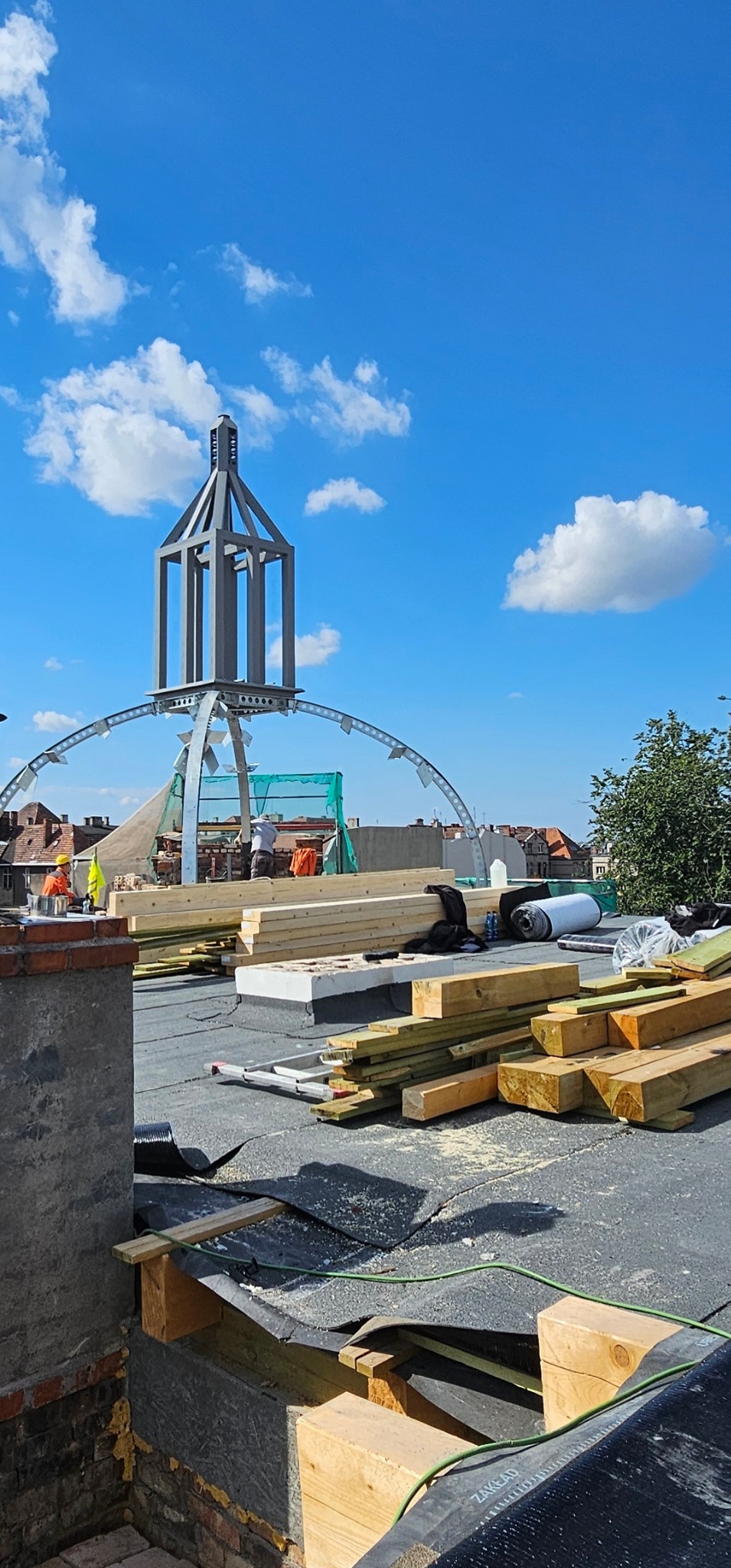 Wysoka konstrukcja na dachu w trakcie budowy, widoczne drewniane elementy, materiały izolacyjne i pracownicy w oddali, błękitne niebo z chmurami w tle.