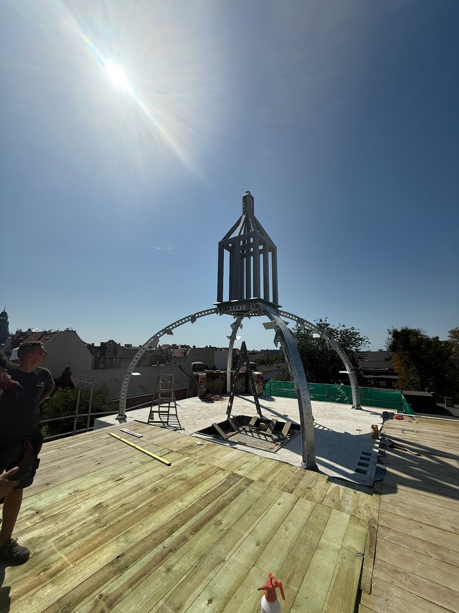 Konstrukcja metalowa na dachu, prawdopodobnie element dekoracyjny lub podstawa pod kopułę. Widoczna drewniana podłoga dachu w trakcie budowy i panorama miasta w tle.
