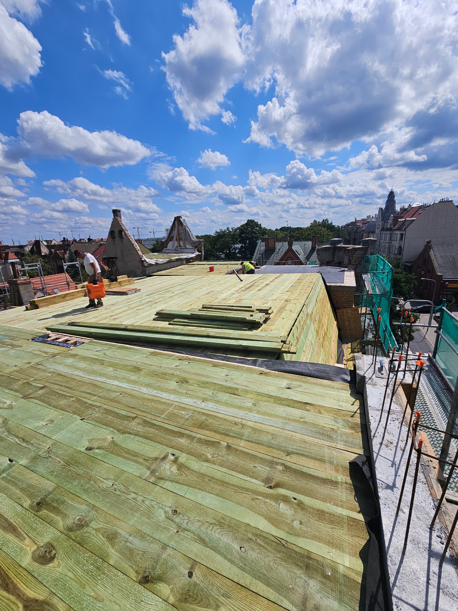 Montaż drewnianej konstrukcji dachu na tle błękitnego nieba z obłokami. Widoczni dekarze podczas pracy. Ujęcie z góry, ukazujące postęp prac.