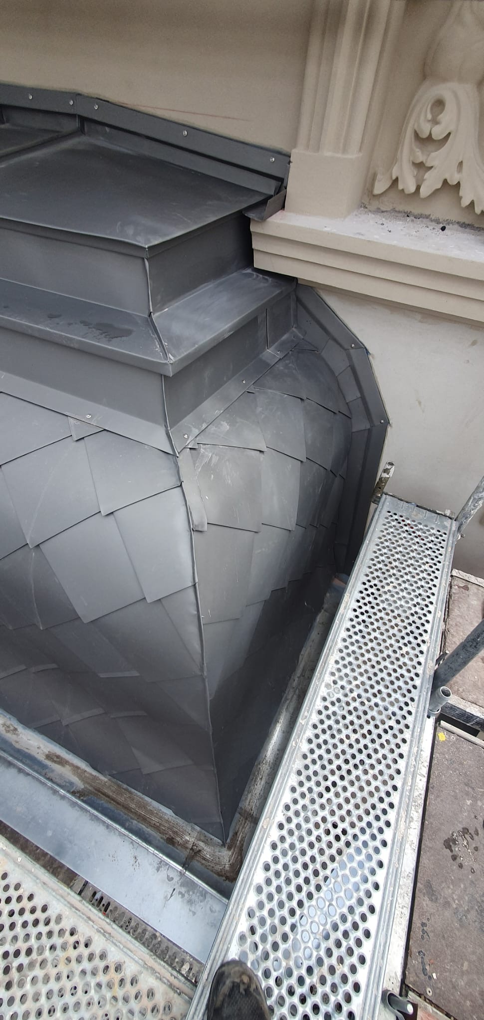 Szary, metalowy dach z łupków w kształcie kopuły z detalami architektonicznymi i fragmentem rusztowania, widziany z bliska pod kątem.