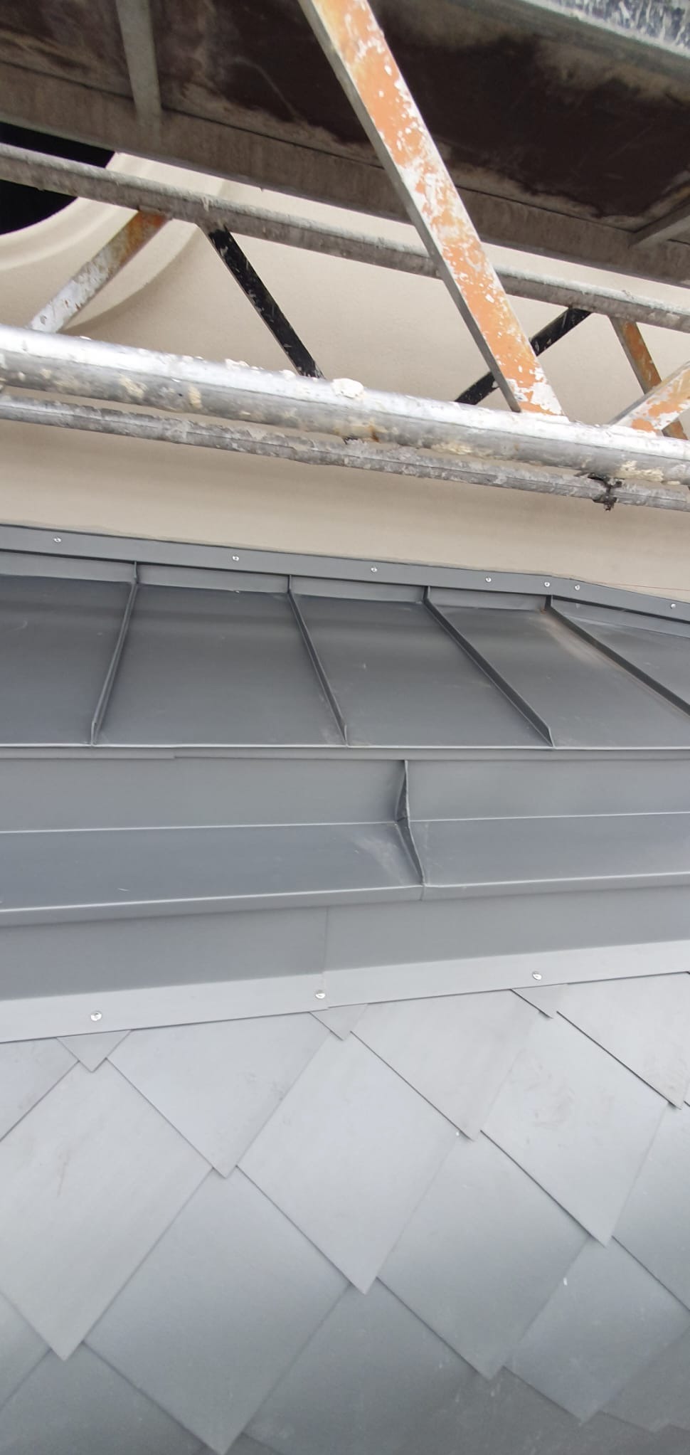 Szare, metalowe płytki dachowe ułożone w romby, widoczne spod rusztowania. Detal dachu z blachy na budynku.